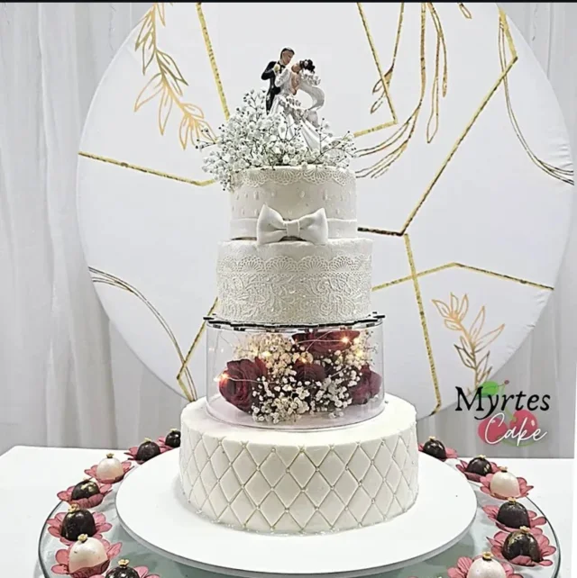 Bolo branco de andares com laço, detalhes de renda, flores e topo com casal, sobre uma mesa decorada.